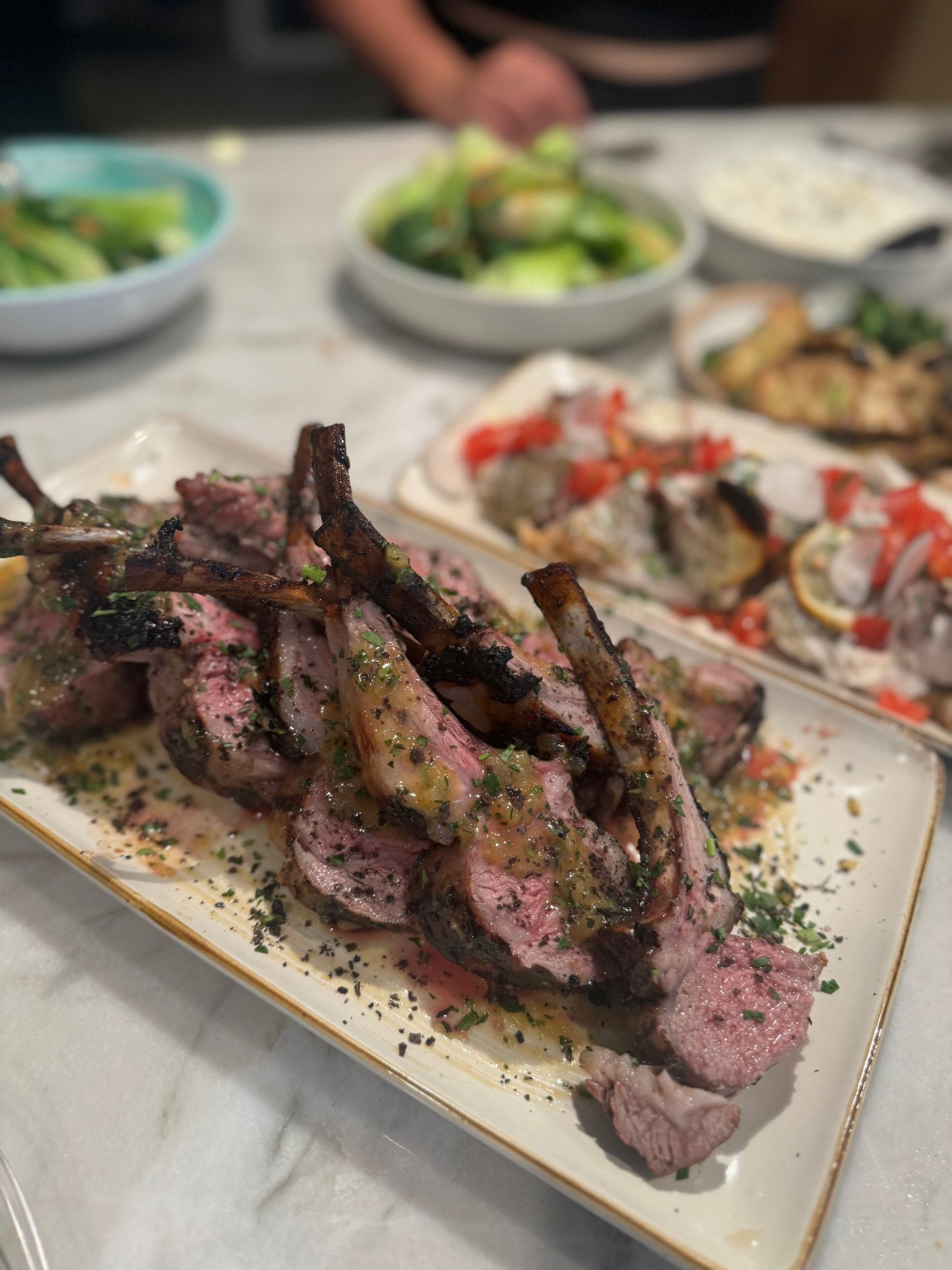 Plated sliced steak with herbs on a table, with salads and other dishes blurred in the background