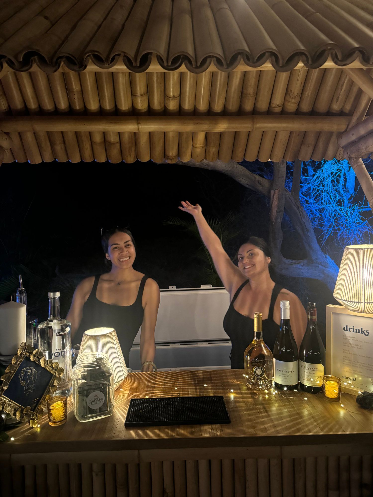 Two people at a bamboo bar at night, with bottles and lanterns on the counter.