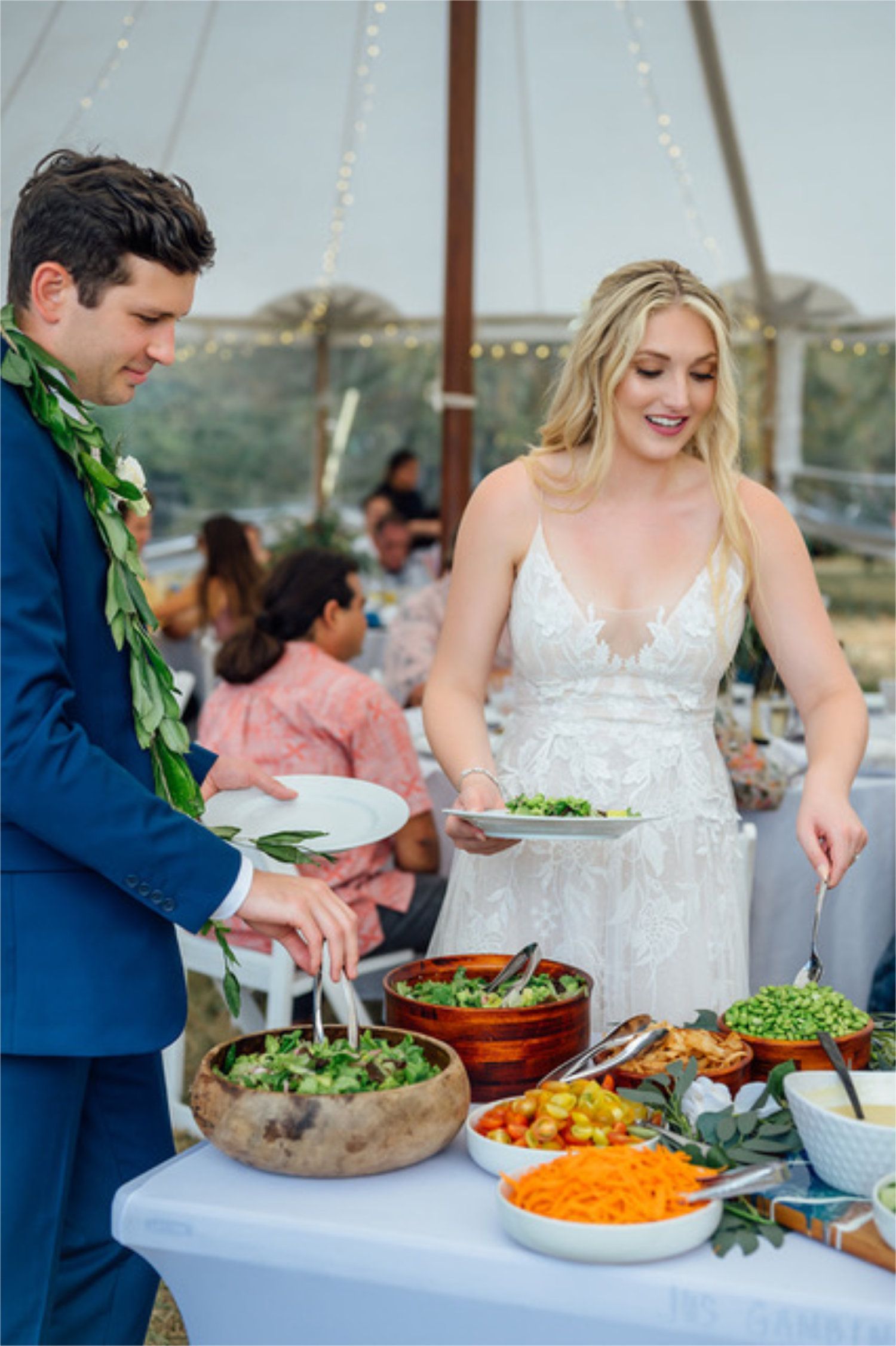 Bride and man serving food at an outdoor buffet with bowls of salad and fruit on a decorated table