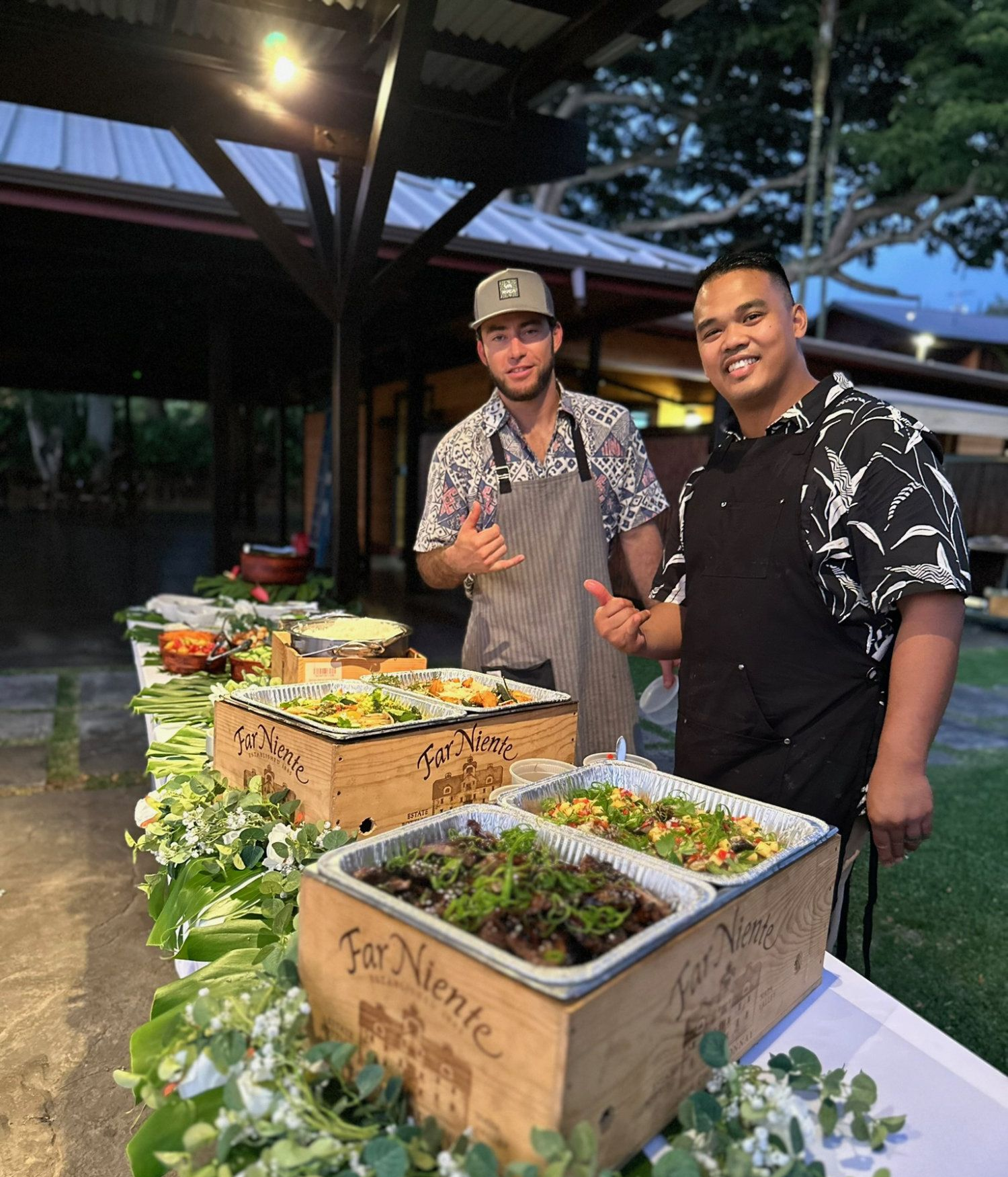 Two smiling people standing behind a buffet table with salads and flowers at an outdoor event