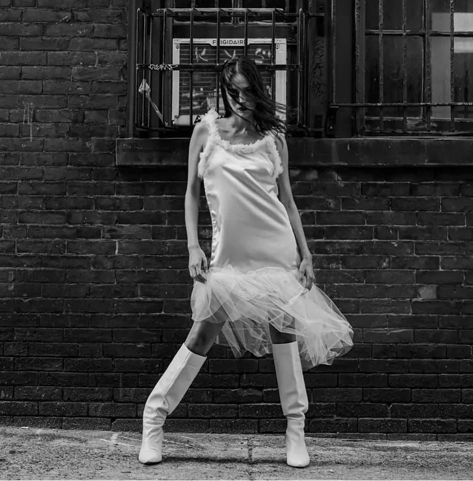 Kylee in a white dress and white boots is standing in front of a brick wall.
