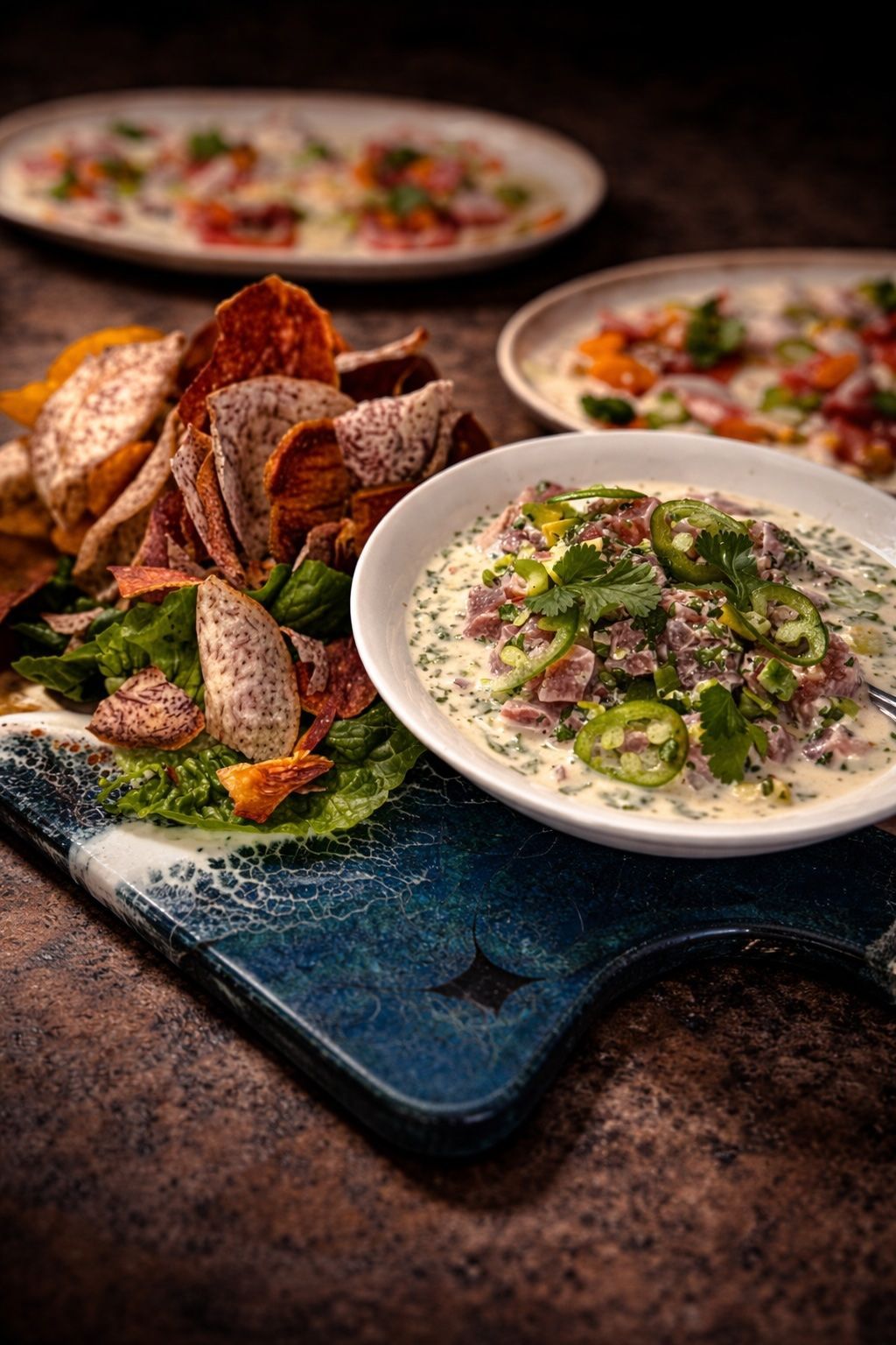Plated gourmet dish with creamy sauce, herbs, and crisp bread on a dark serving board, with two blurred plates behind