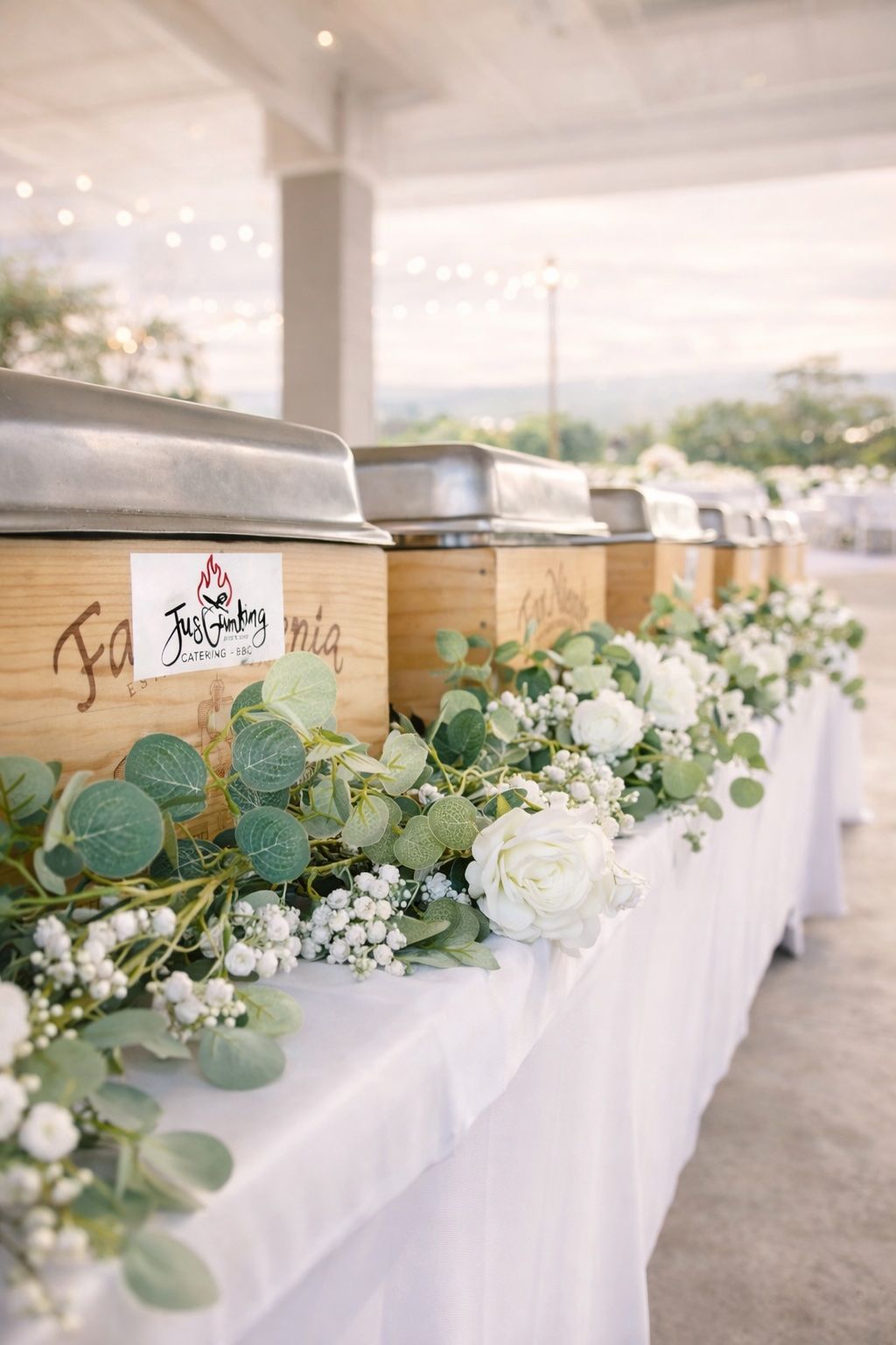 Row of wooden beverage dispensers decorated with white flowers and greenery at an outdoor event