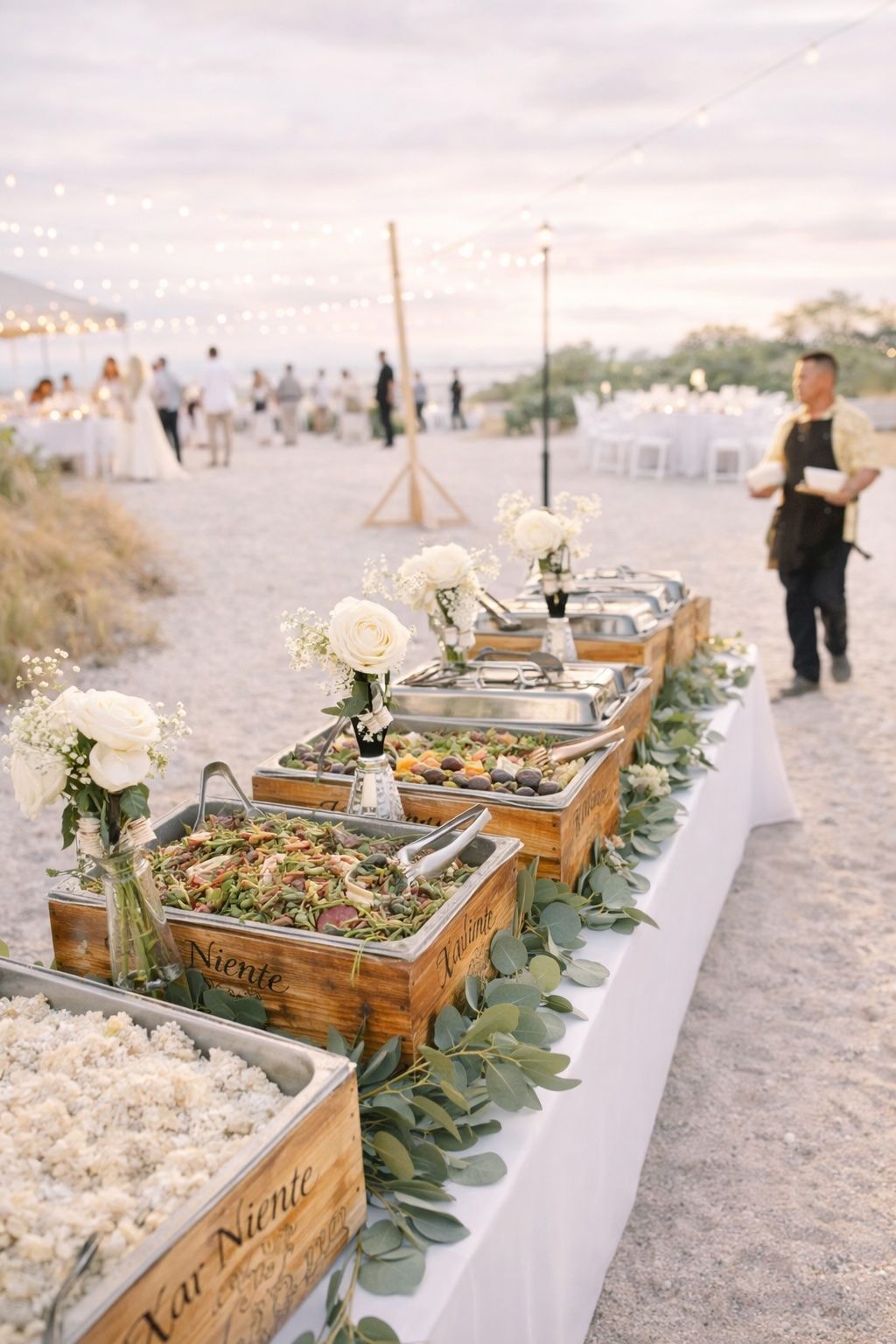 Outdoor buffet table with white flowers and greenery at a sunlit event, with a person standing nearby
