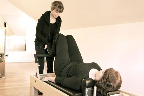 A woman is laying on a pilates reformer while another woman stands behind her.