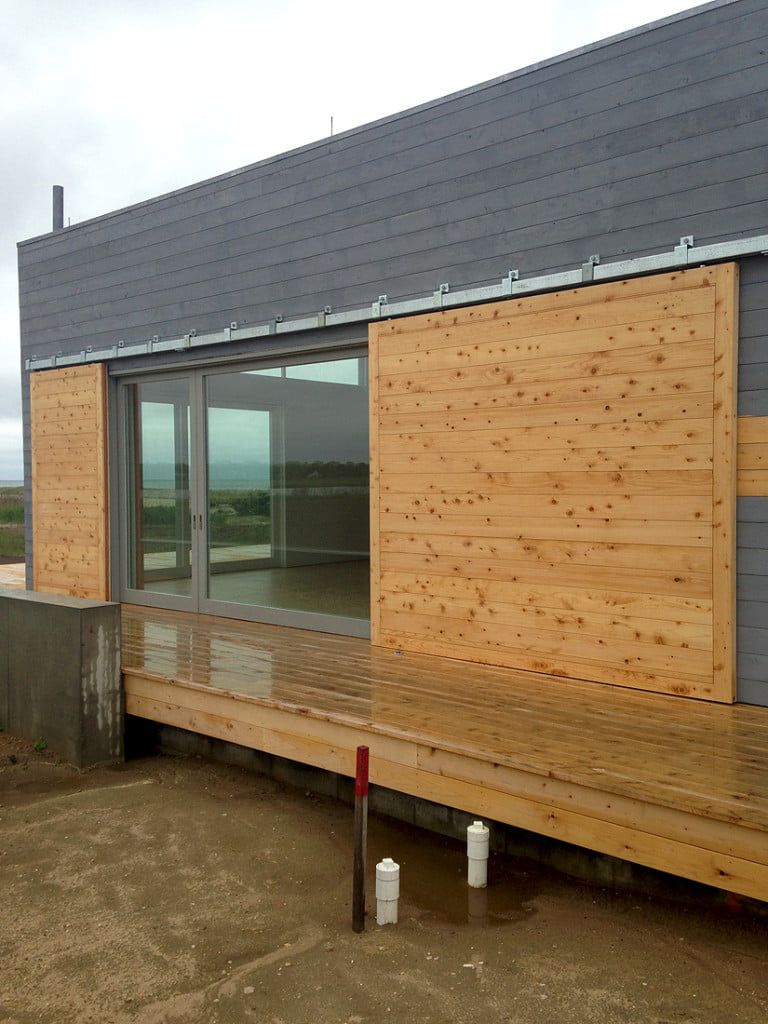 Modern house exterior with plywood-covered windows, large sliding glass door, and gray roof.