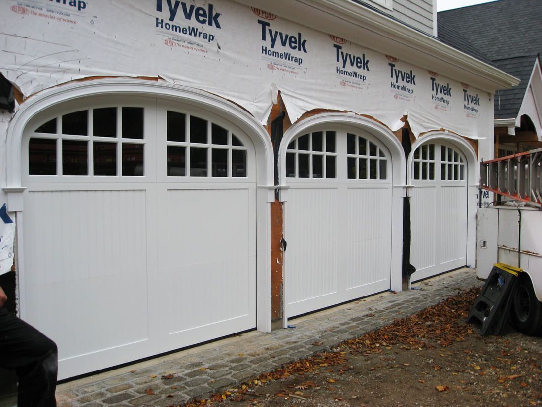 Three white garage doors with arched windows on a house under construction, wrapped in Tyvek.