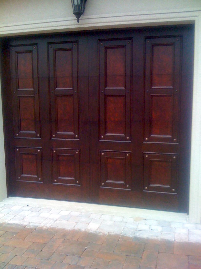 Dark wooden double garage doors on a brick driveway under a wall lamp