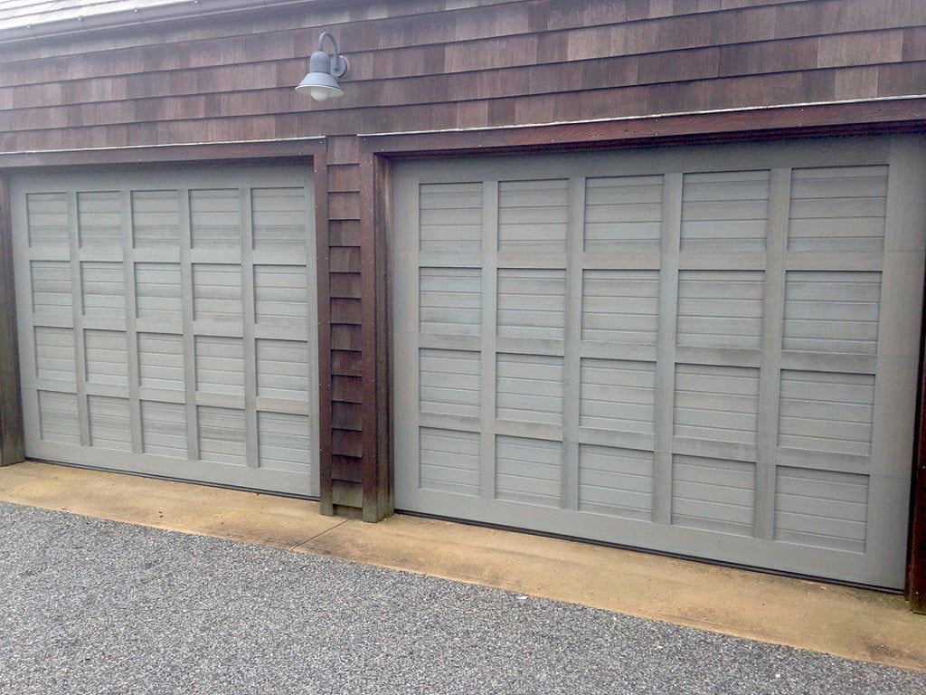 Two gray garage doors on a brown shingled building with a gravel driveway and wall light above