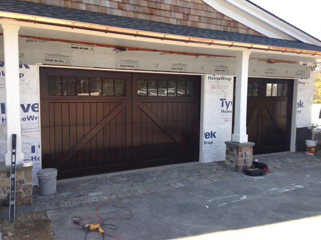 Two dark brown garage doors on a house under construction, with exposed siding and tools on the driveway.