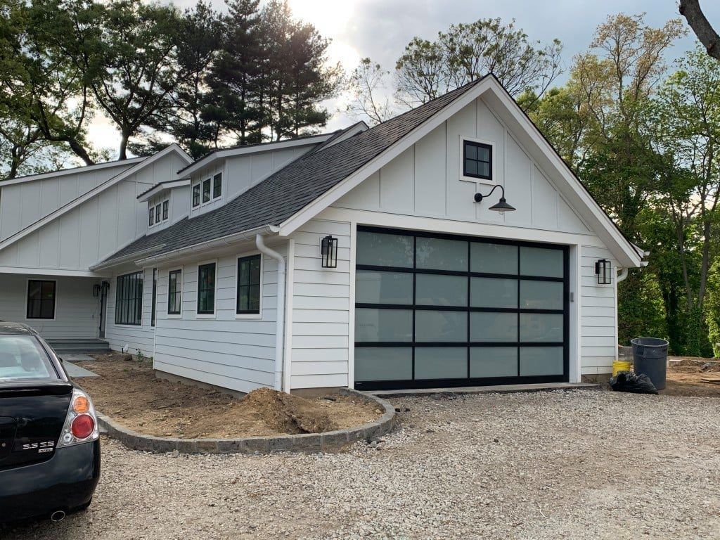White house with gray roof and large garage door beside a gravel driveway and parked car.