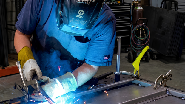 Welder in blue shirt and helmet welding metal on a workbench, bright sparks flying.