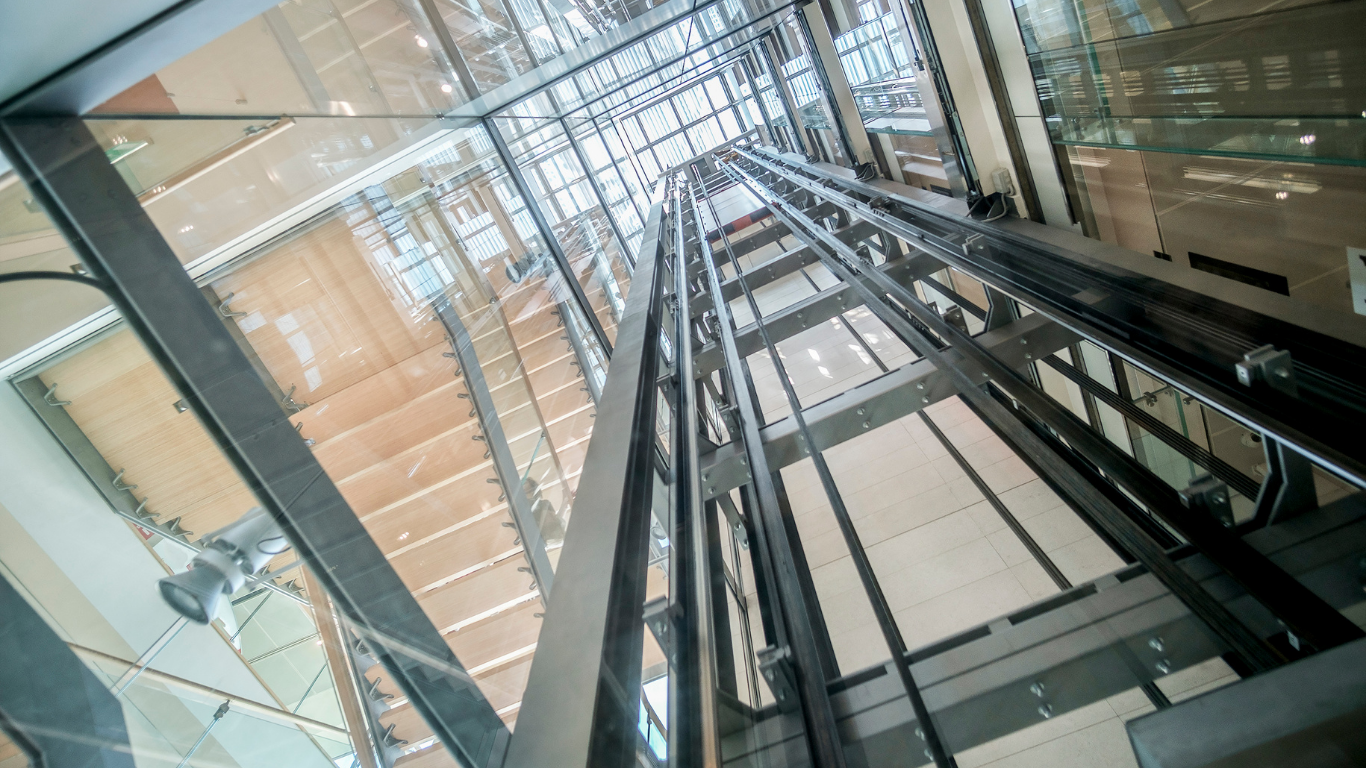 Looking up inside a glass elevator shaft with metal beams and reflections in a modern building.