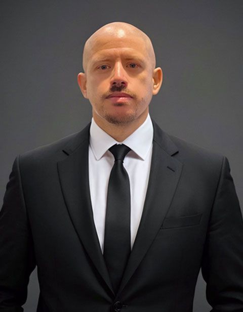 Hombre con traje negro y corbata, calvo, expresión seria. De pie frente a un fondo gris.