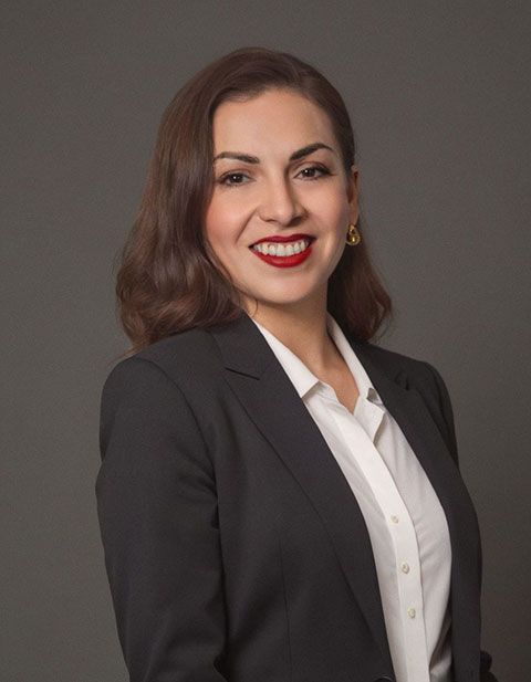 Mujer con blazer negro y camisa blanca, sonriendo, sobre un fondo gris.