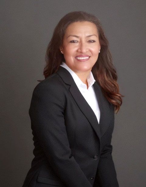 Mujer con blazer negro y camisa blanca, sonriendo, sobre un fondo gris.