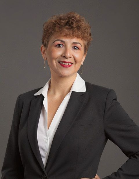 Una mujer con un traje negro sonríe, con los brazos a los costados y una camisa blanca con cuello debajo.