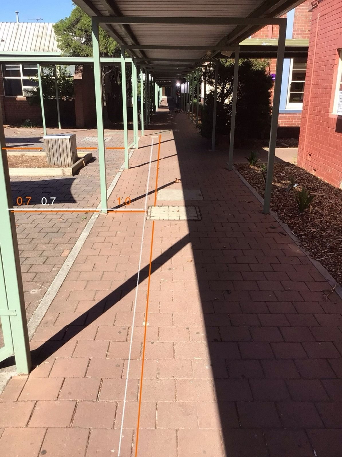 A long, brick walkway shaded by a green metal awning. Strong shadows cast by the sun.
