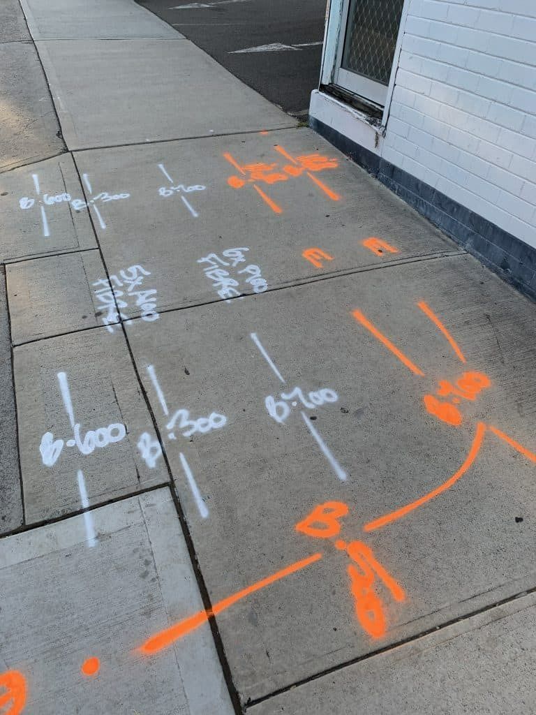 Sidewalk with white and orange markings for utility work, near a building.