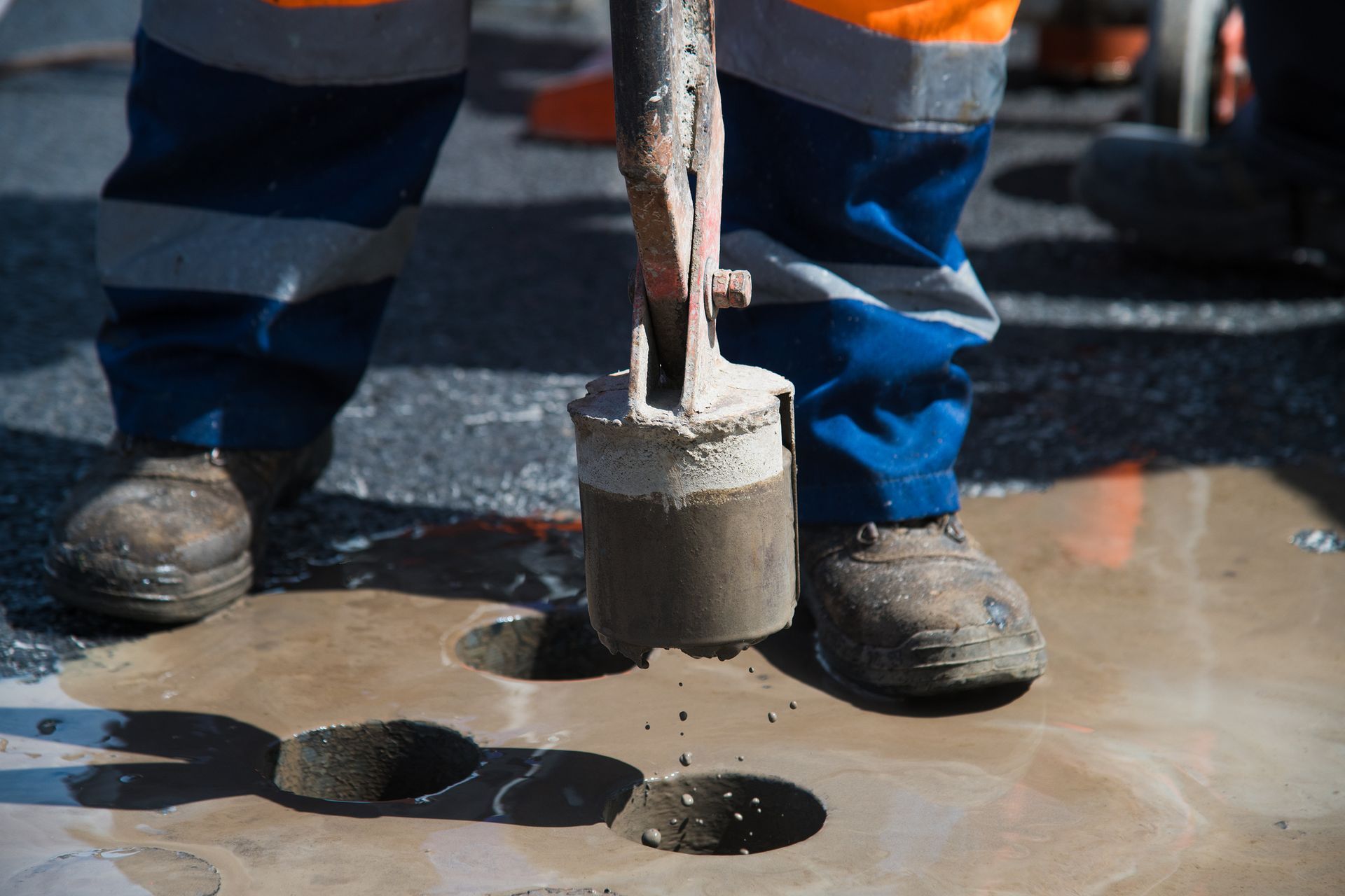 Person in work clothes using a core drill to bore into a paved surface.