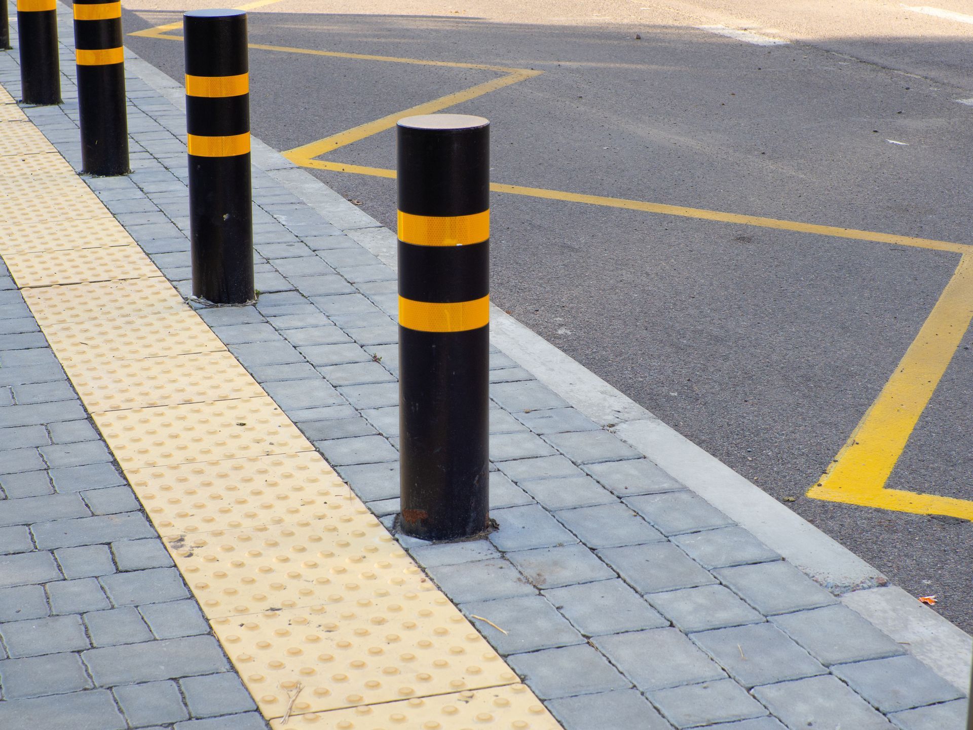 Black bollards with yellow stripes line a sidewalk with tactile paving next to a street.