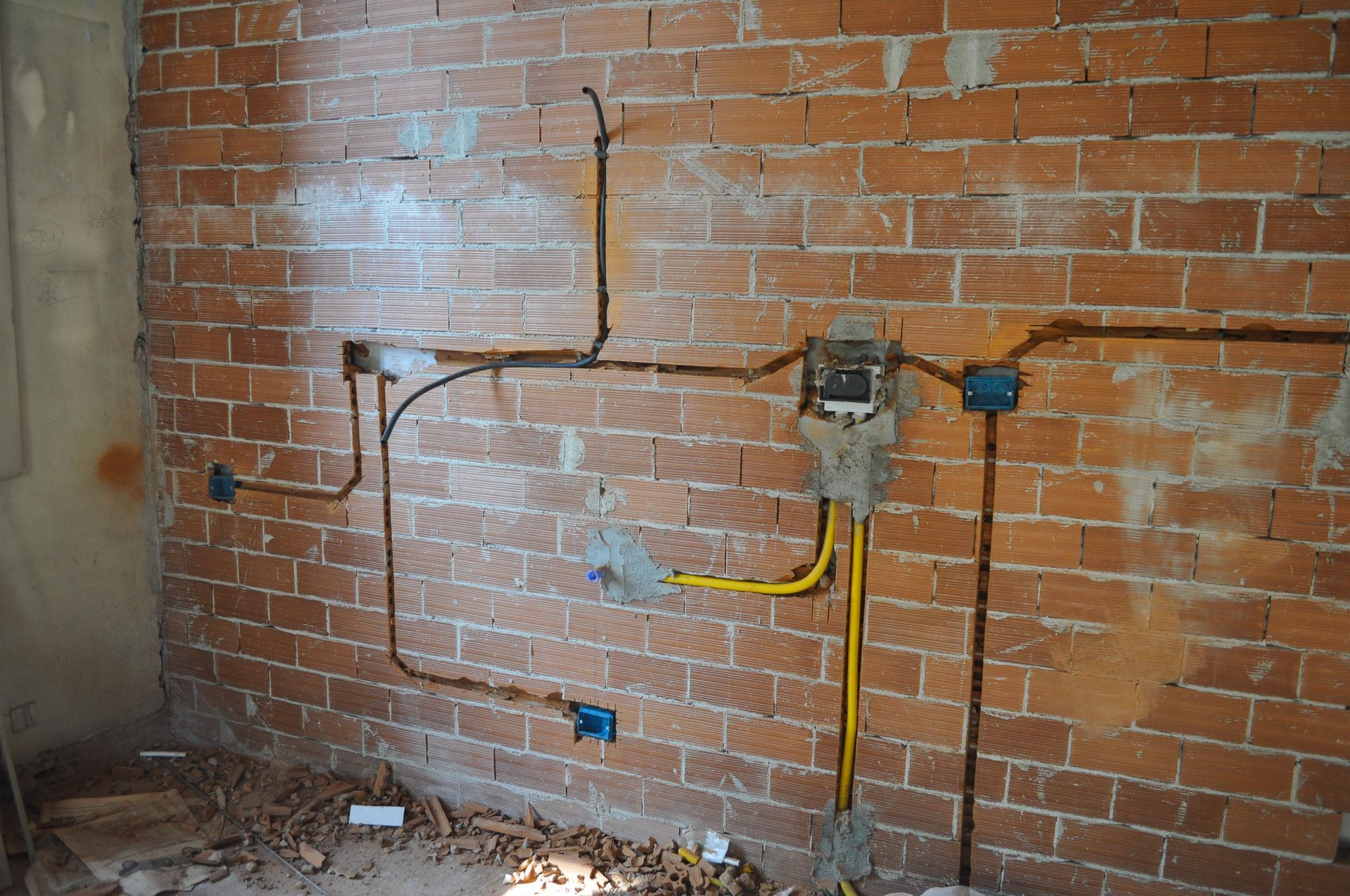 Electrical wiring on a brick wall with conduit, junction boxes, and a light-colored ground covering.