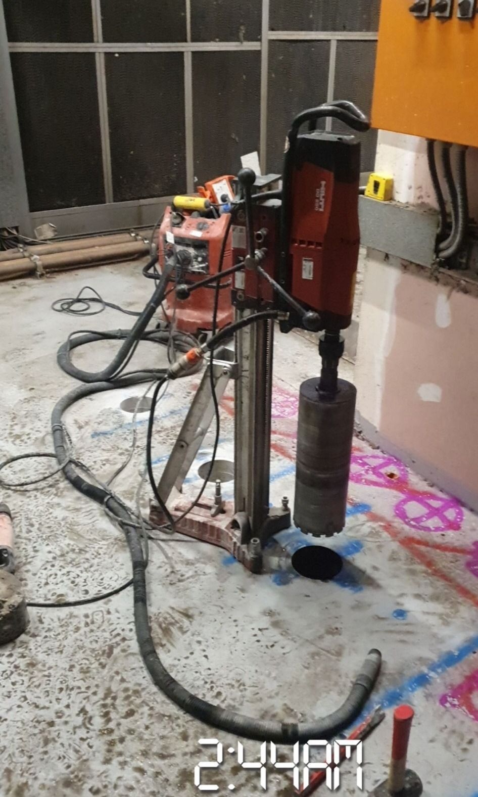 A core drilling machine on a concrete floor is drilling a hole. The setting appears to be an unfinished building.