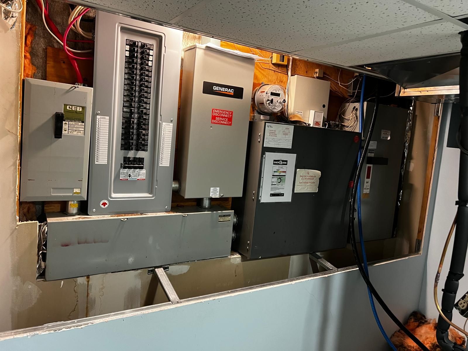 Electrical panel installation in a basement, with various grey boxes and wires against a wall.
