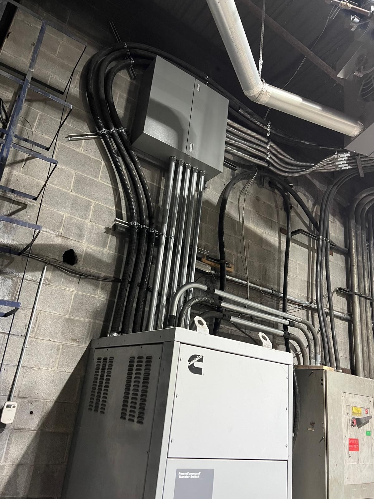 Electrical equipment on a gray cinder block wall, including conduits, a Cummins unit, and junction boxes.