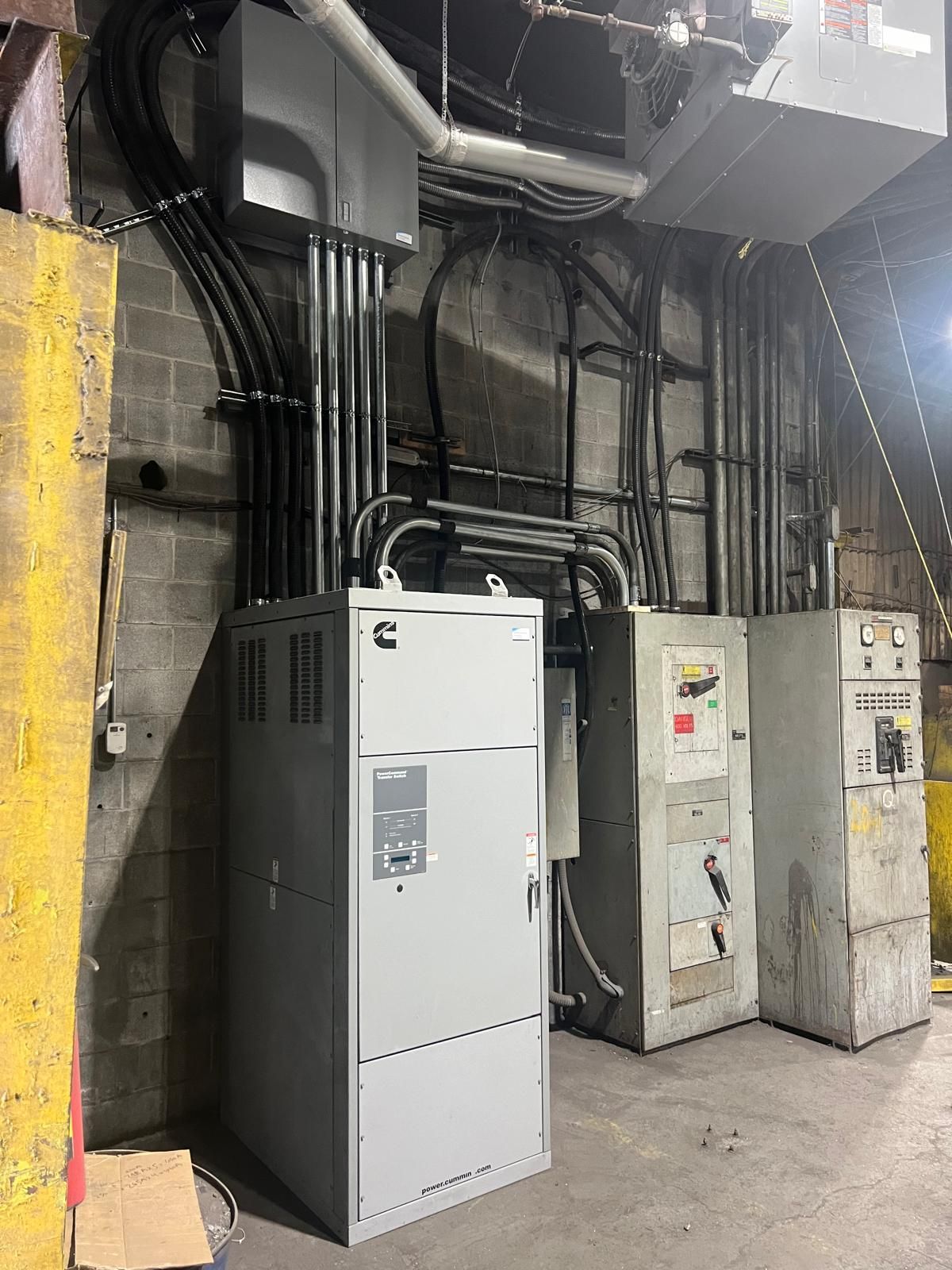 Electrical equipment in a commercial space, including a large gray GE panel, other metal boxes, and conduit pipes against a brick wall.