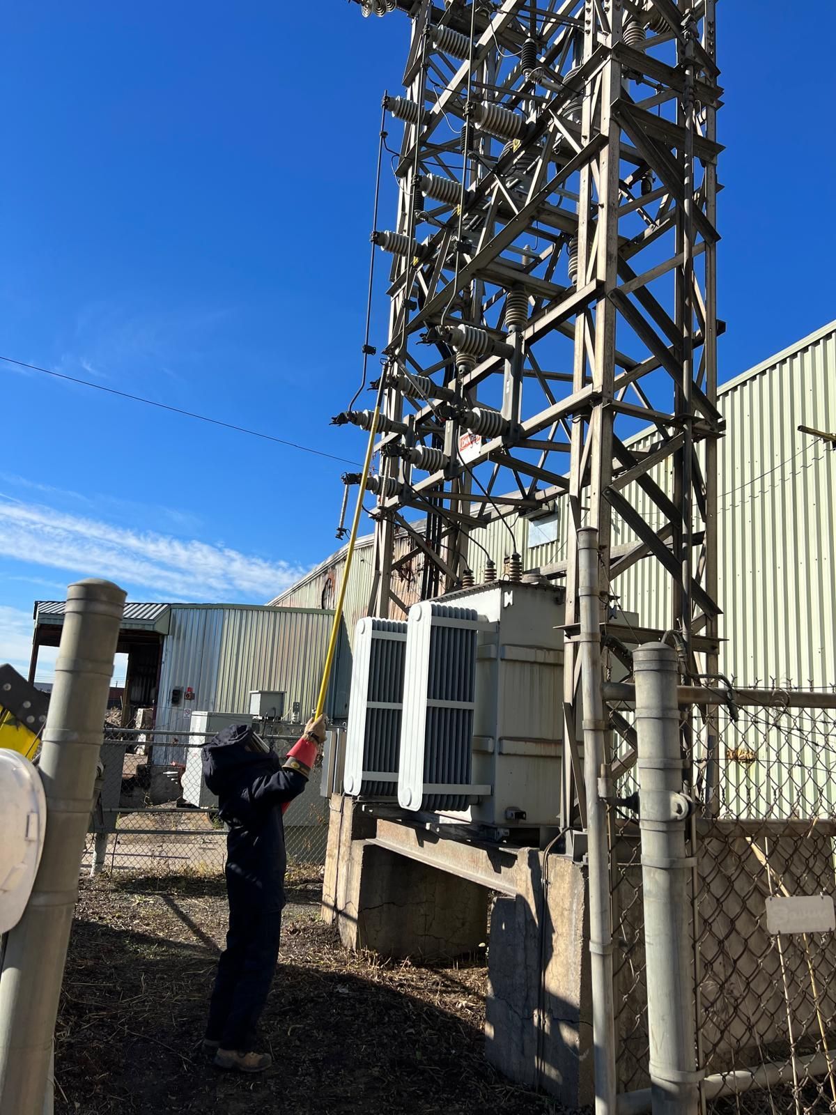 A person measures a utility transformer next to a metal tower and a building on a sunny day.