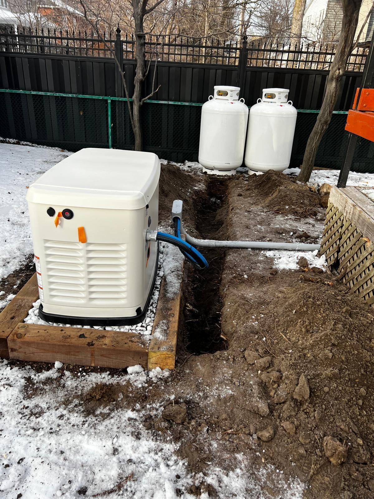 A generator next to propane tanks in a backyard, with a trench and a snow-covered ground.