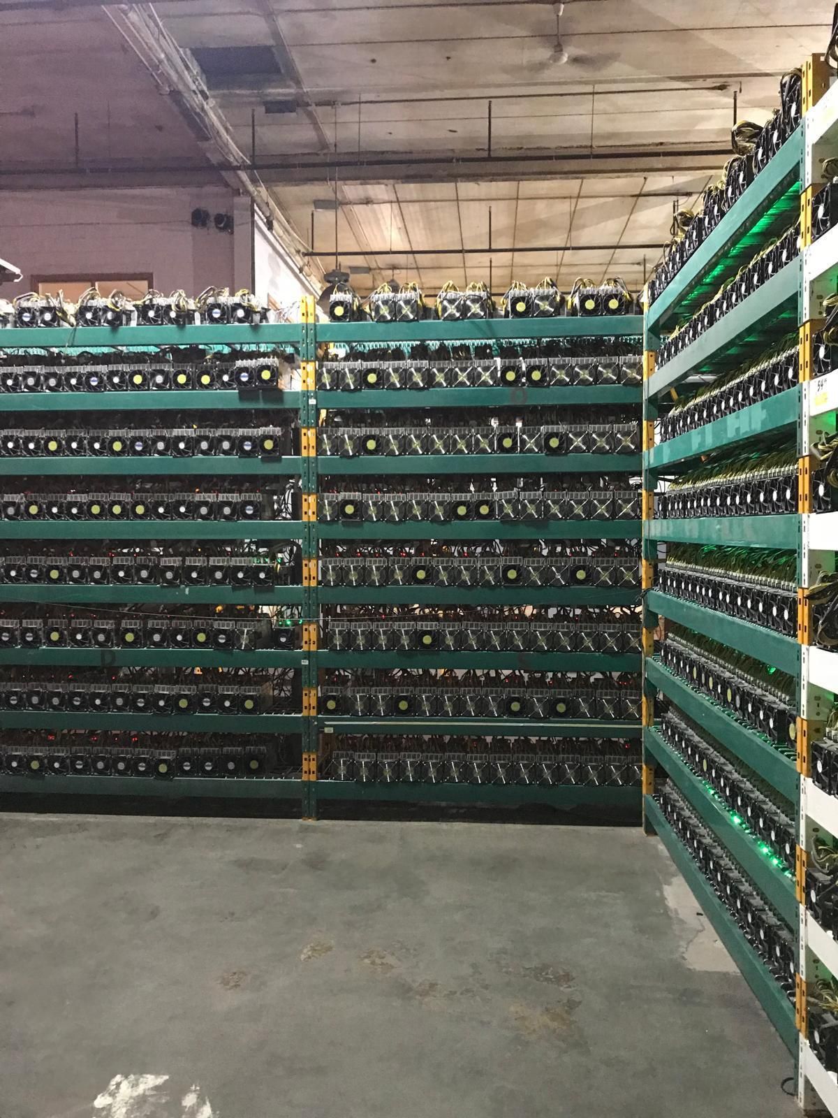 Rows of cryptocurrency mining rigs in a warehouse setting; dark green racks filled with computer hardware, visible ventilation fans.