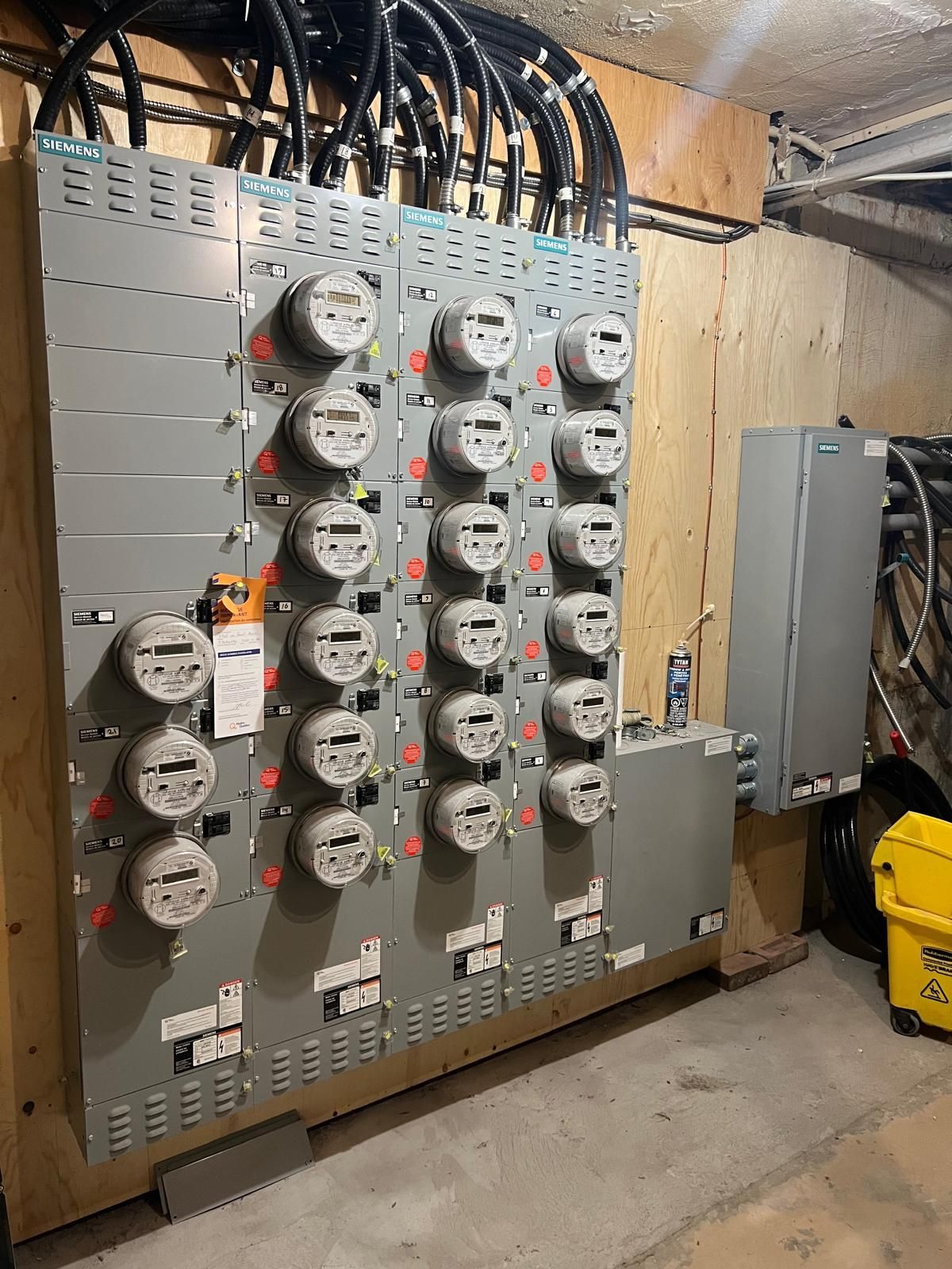 A row of electrical meters in a building, mounted on a gray metal panel against a wood wall. Black cables lead to the meters.
