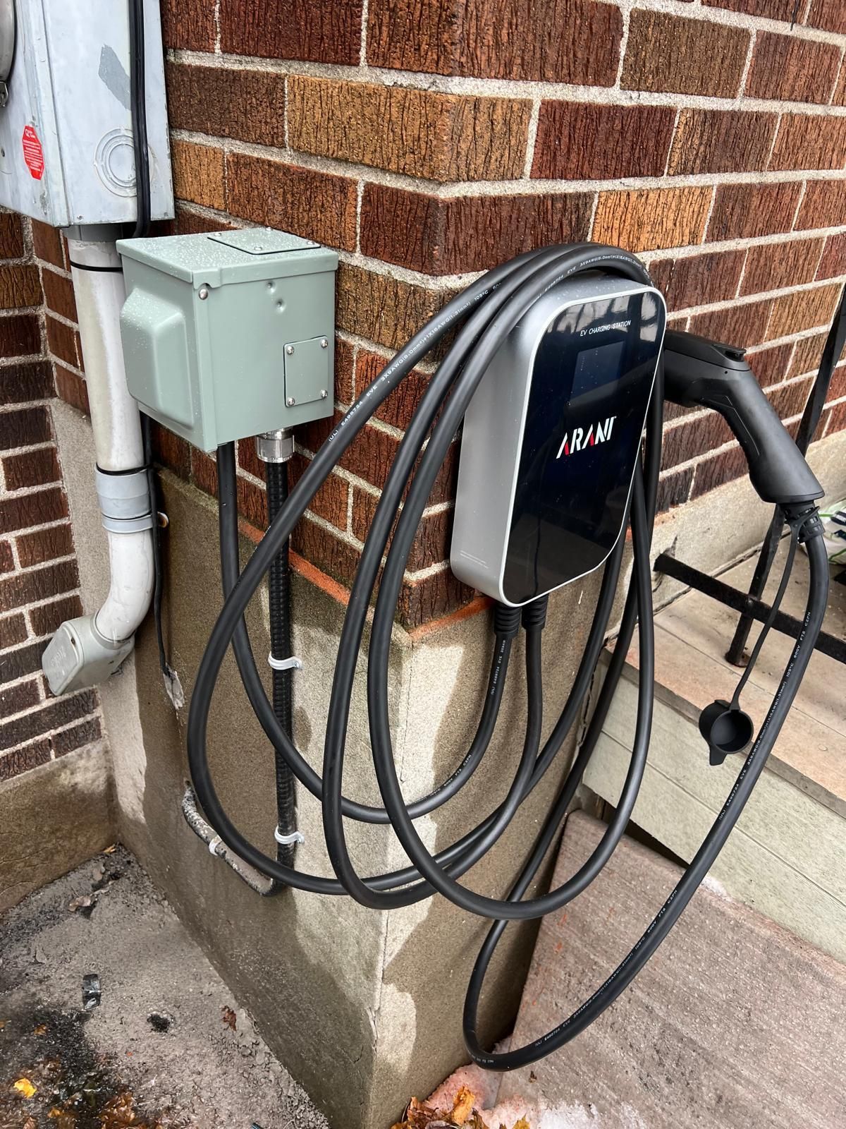 Electric vehicle charger mounted on a brick wall with its cable coiled. A gray electrical box and conduit are adjacent.