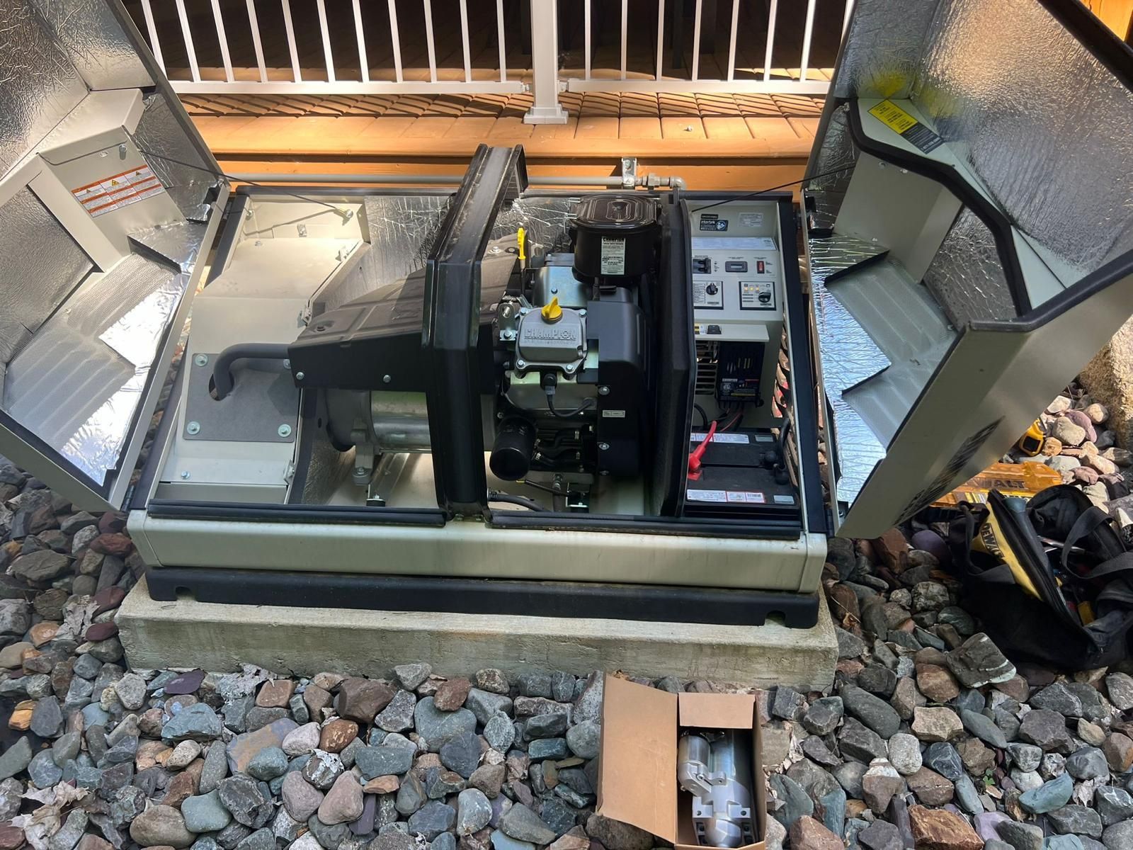 A standby generator with open panels sits on a concrete pad surrounded by gravel. The unit is gray and has a visible engine and control panel.