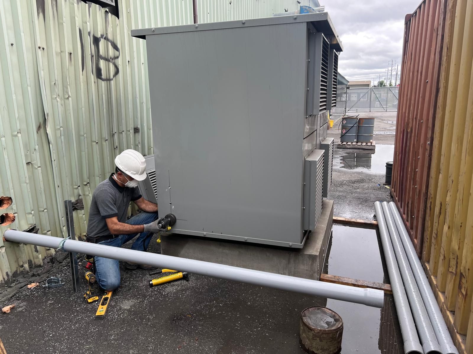 A worker in a hard hat and face mask installs electrical conduit alongside a large gray utility box, outdoors between walls.
