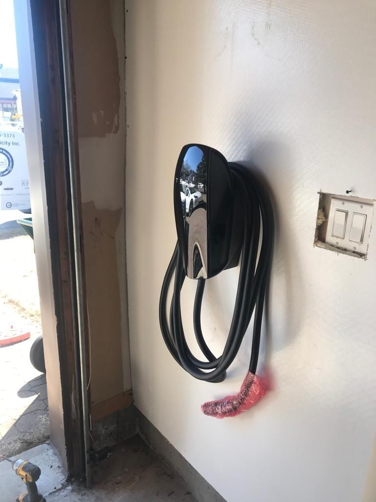 Black electric vehicle charger mounted on a white garage wall, cord coiled neatly. Nearby is an open electrical outlet.