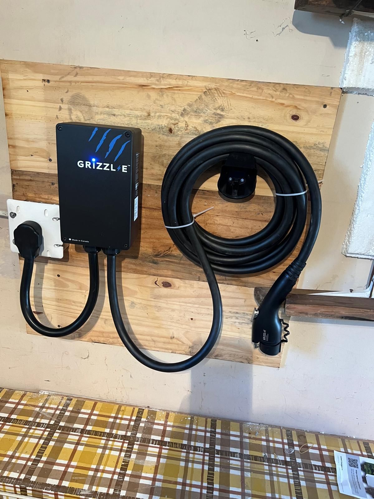 An EV charger mounted on wood. Black charger and coiled cable. Plugged into a white outlet.