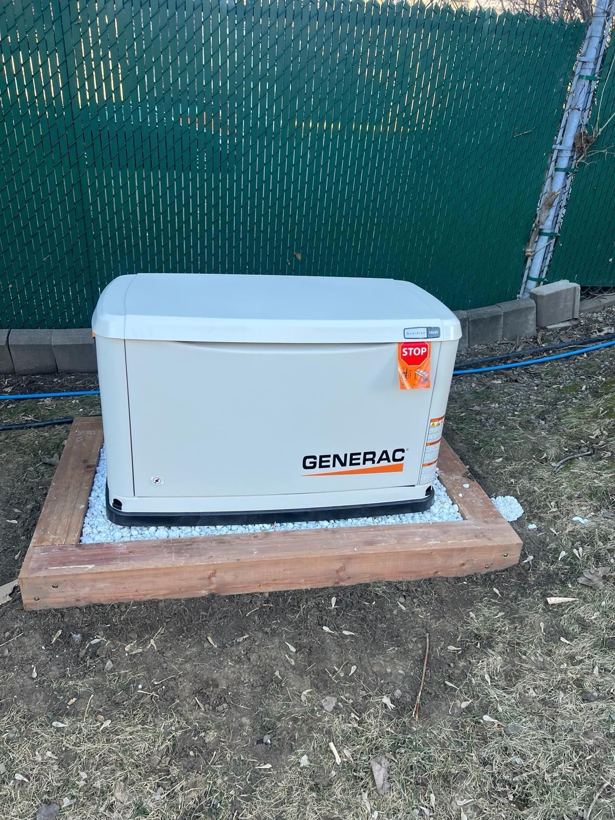 Un générateur Generac beige sur une plate-forme en bois dans une cour avec une clôture verte.