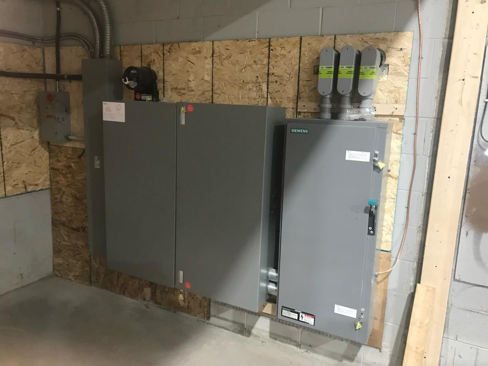 Electrical panel and boxes mounted on wood paneling; conduits and wires visible; gray and neutral colors.