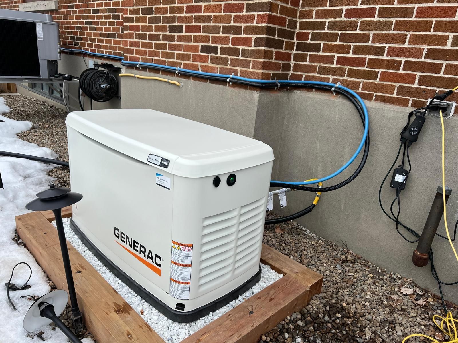 Un générateur Generac repose sur une plateforme en bois à côté d'un bâtiment en briques. Des fils électriques noirs et bleus en partent.