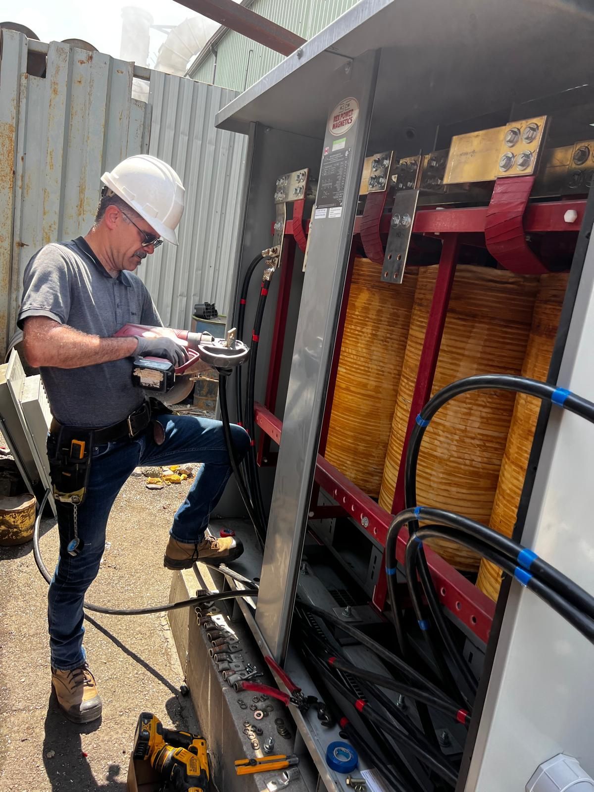 A worker in a hard hat and work clothes inspects electrical equipment inside a large, open metal cabinet outdoors.
