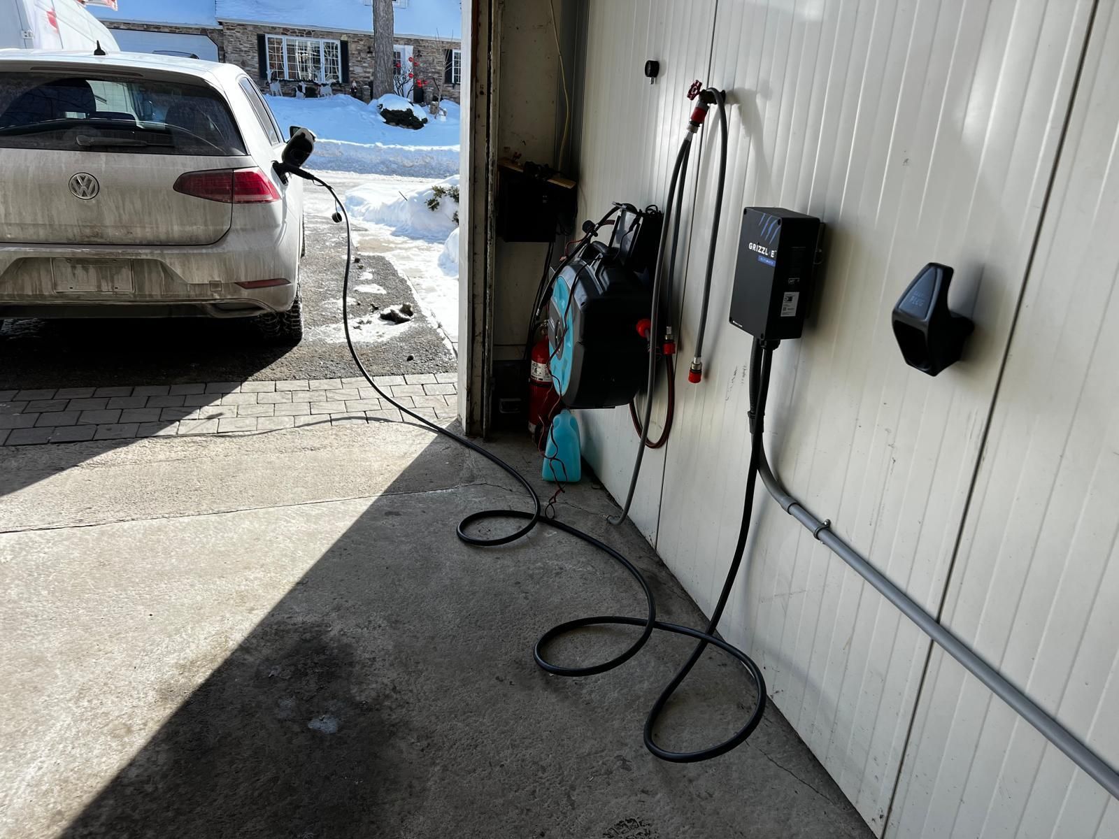 White car charging in a garage; charging cable connected. Snow on the ground, sunny day.