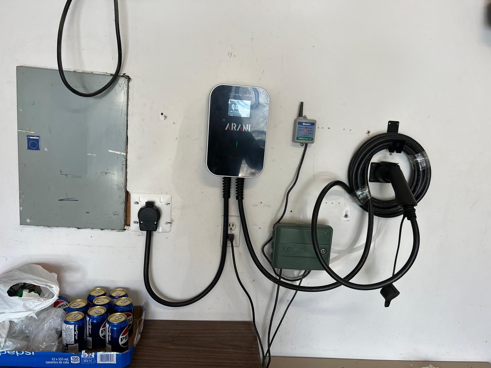 An electric vehicle charger mounted on a white post. The charger is black with a green light, with a black cord hanging down.