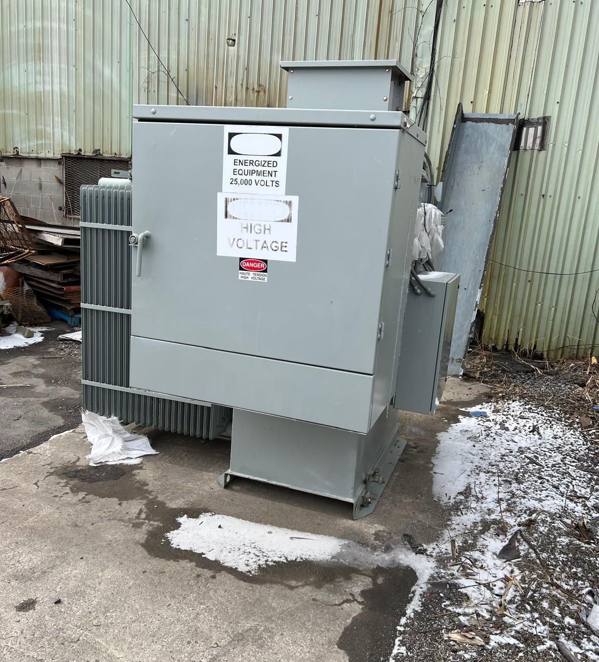 Gray electrical transformer on concrete, with high voltage warning signs. Set outdoors near a corrugated metal wall.