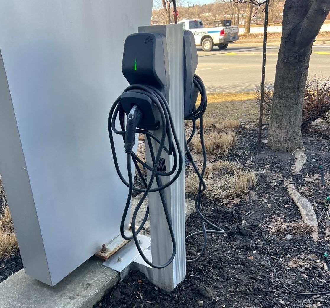 An electric vehicle charging station outdoors, mounted on a post next to a concrete structure.  The charging cable hangs down onto the ground.