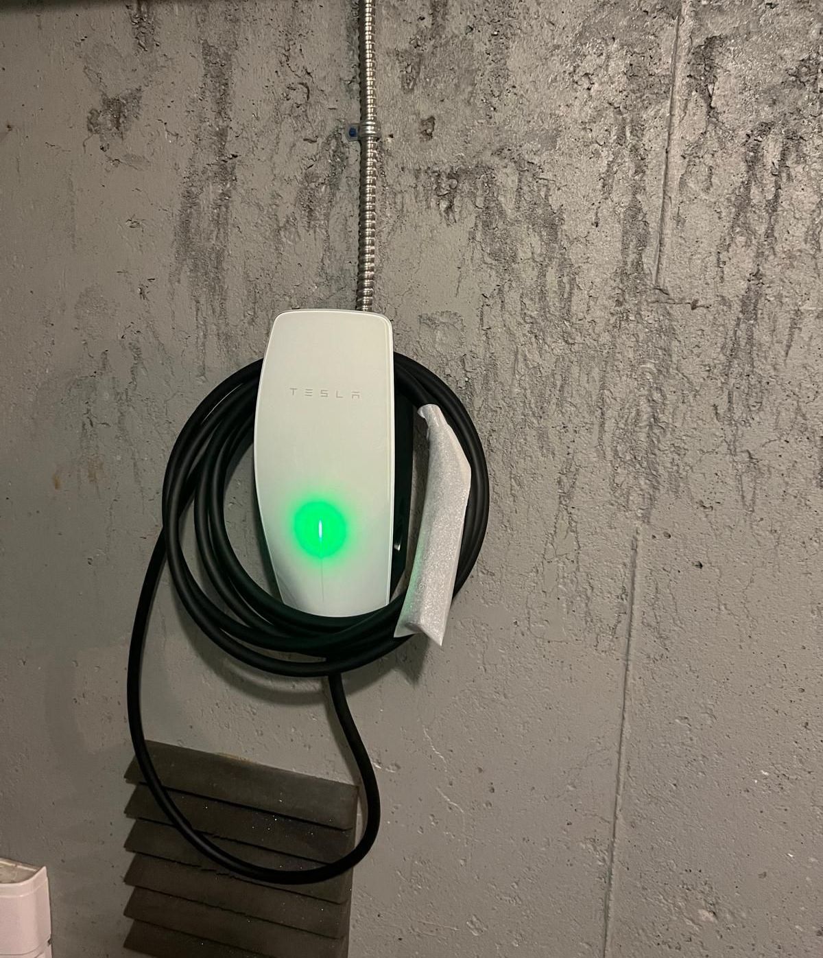 Wall-mounted white EV charger with green light and black cable on a gray concrete wall.