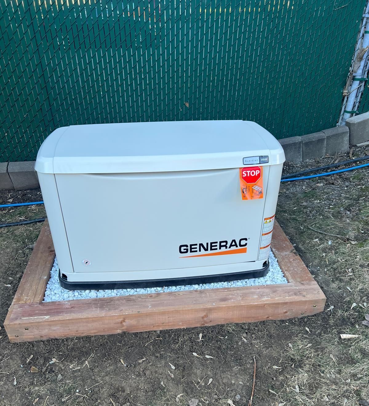 A Generac generator on a wooden platform, against a green fence. It's an off-white color, with the brand name visible.