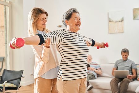A woman is helping an elderly woman lift dumbbells in a living room.